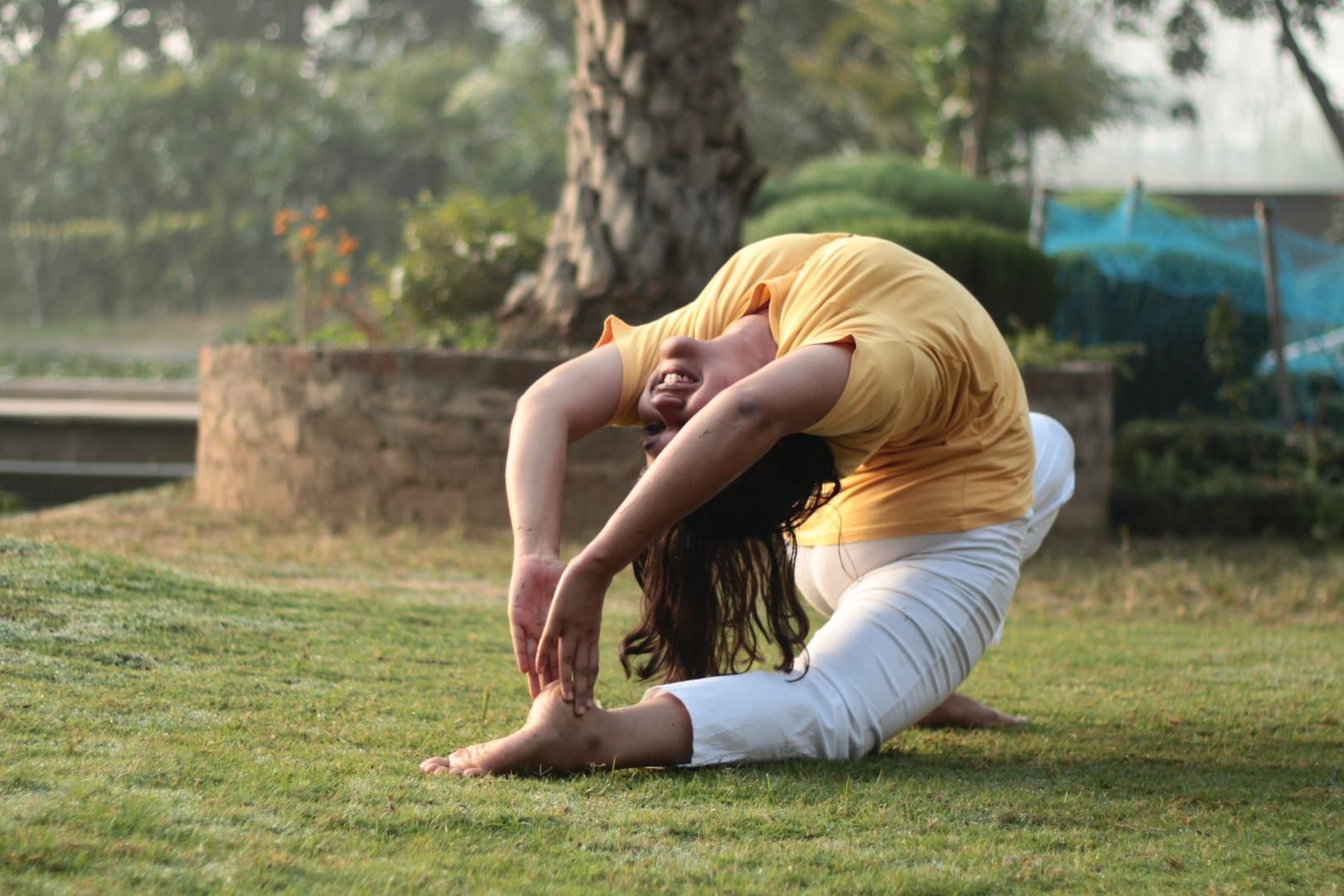 A student in a yellow T-shirt and white trousers kneels in a backbend on grass, reaching both hands toward the feet.