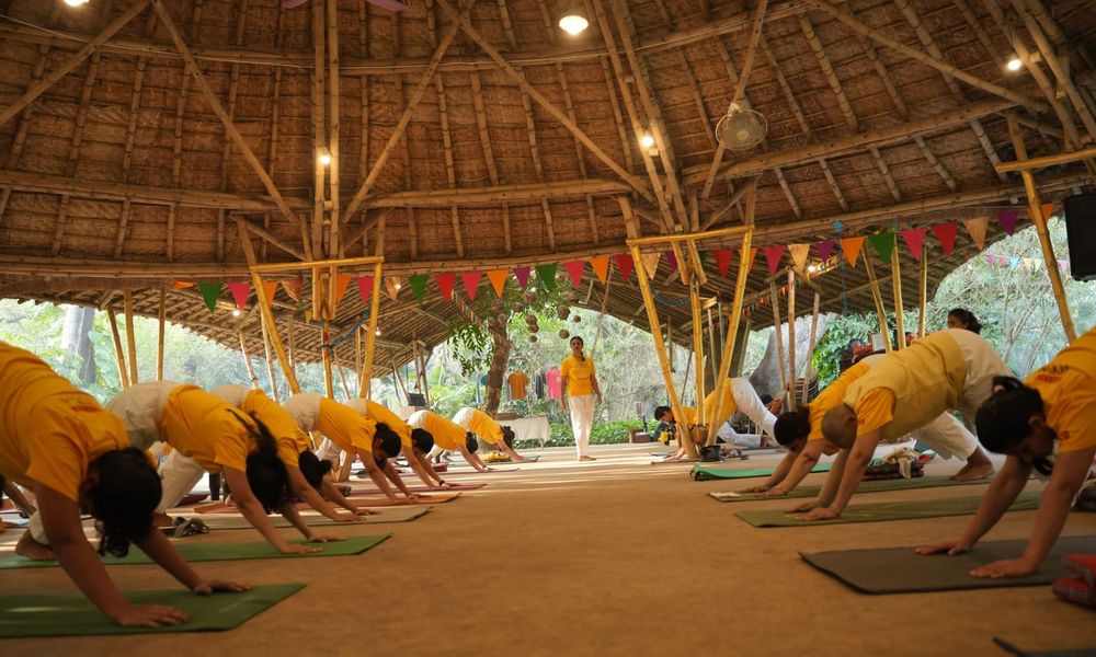 Students holding Adho Mukha Svanasana (downward-facing dog) on their mats as a teacher watches.
