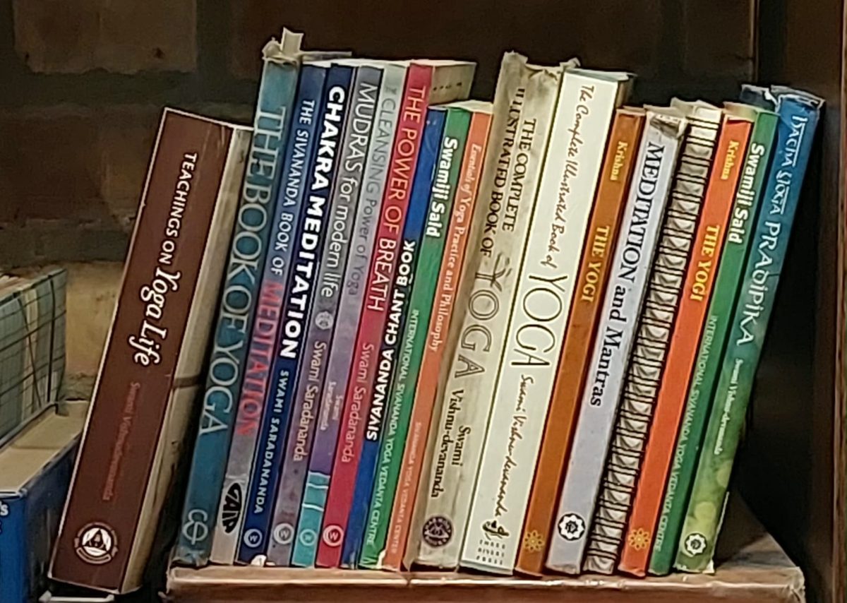 A row of yoga and meditation books standing spine-out on a shelf at Sivananda Gurgaon, among them The Complete Illustrated Book of Yoga; Meditation and Mantras; the Sivananda Chant Book; and Hatha Yoga Pradipika
