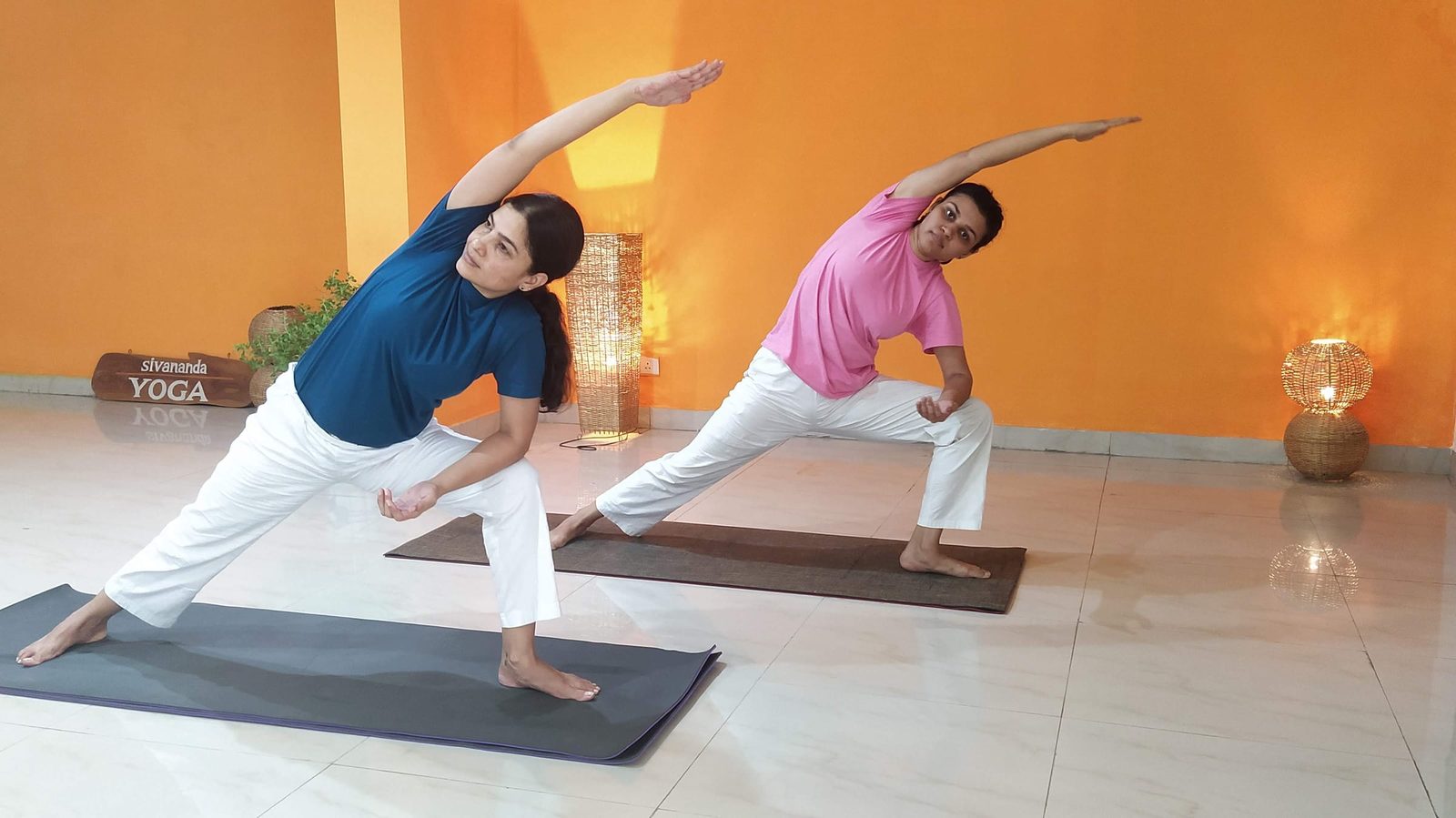 Two students practicing a standing side bend at Sivananda Gurgaon, each with one arm reaching overhead, on mats in front of an orange wall carrying the centre's Sivananda Yoga sign.
