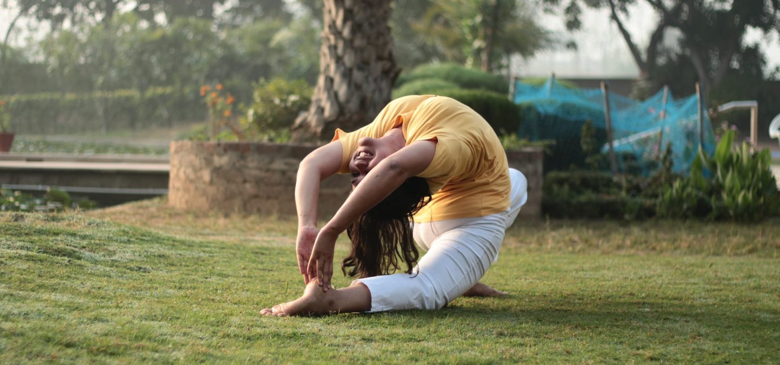 A woman in a yellow T-shirt and white trousers arches into a deep backbend on a lawn, one leg stretched forward and both hands reaching for the front foot