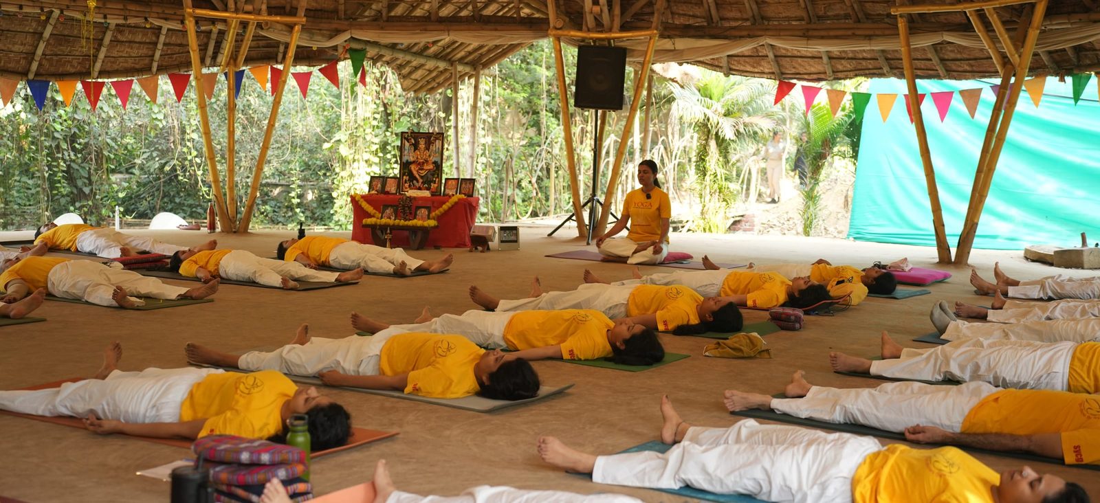 A full class lying in Savasana on mats under the pavilion, a teacher seated before the altar at the far end