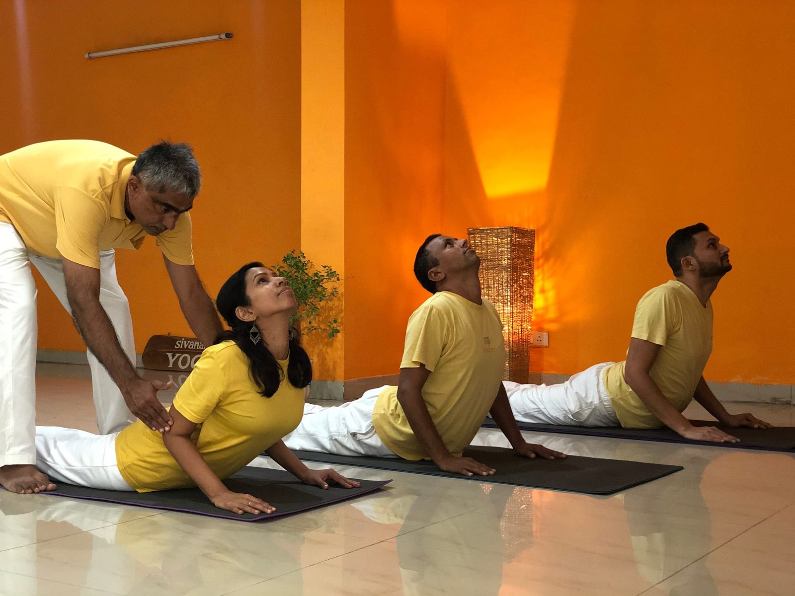 A teacher at Sivananda Gurgaon guiding a student in Bhujangasana (Cobra), with two more students in the pose beside them.