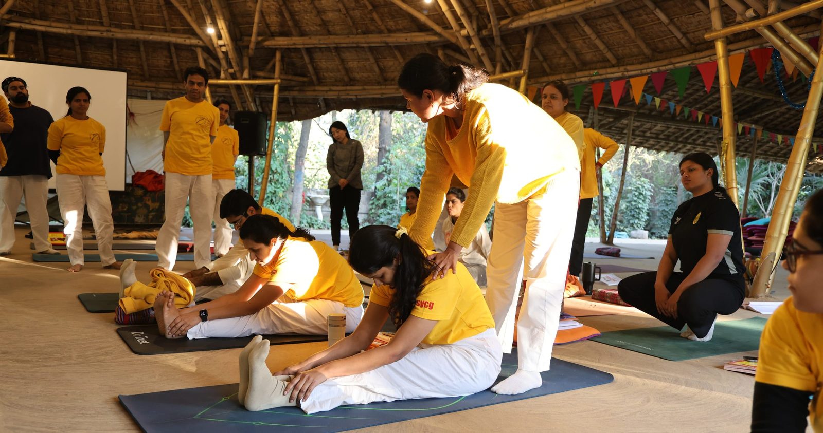 A teacher adjusting a trainee in a seated forward bend during a teacher training course