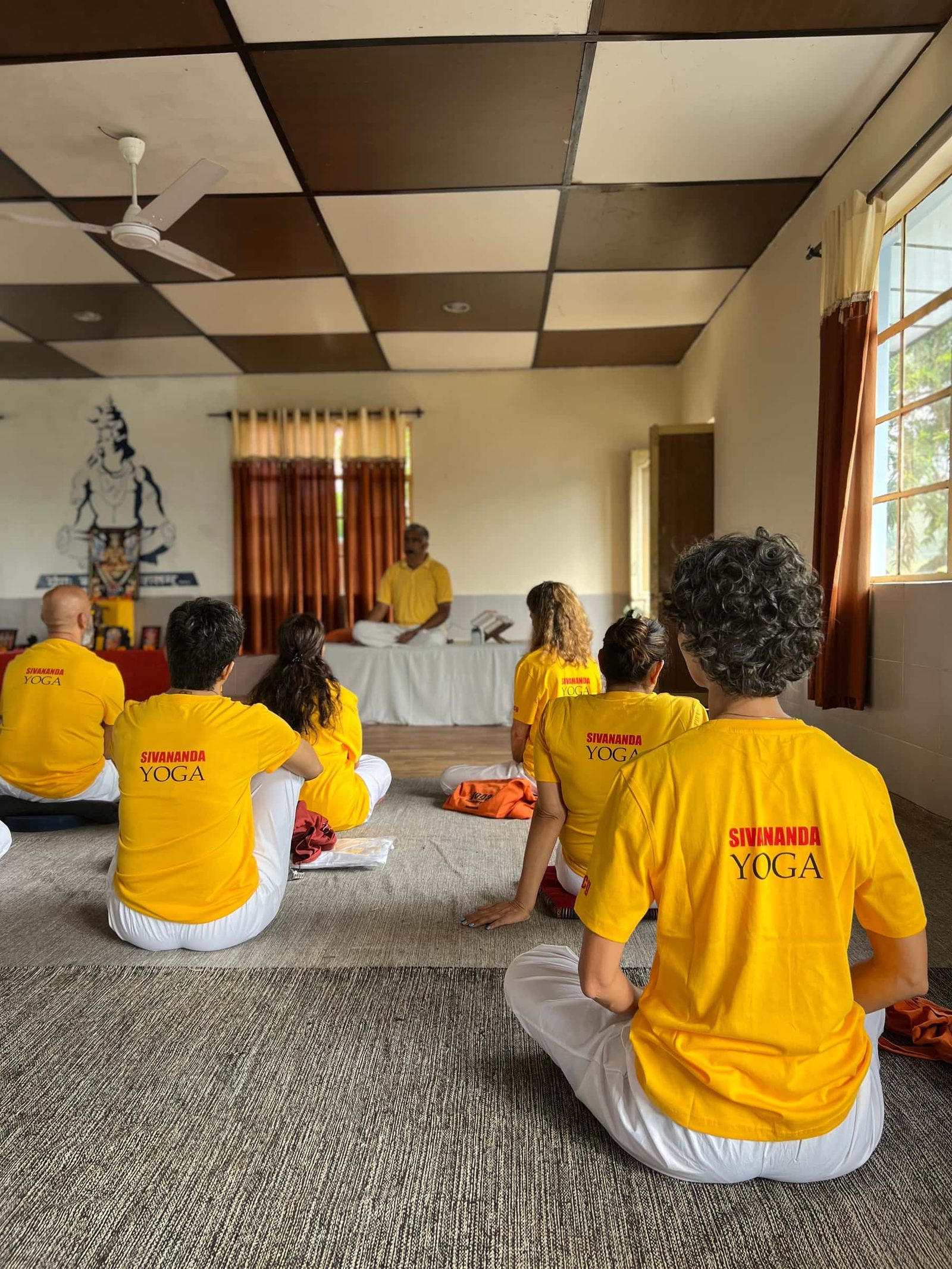Students in Sivananda Yoga shirts seated on the floor facing a teacher on a low platform, a painted mural on the wall behind