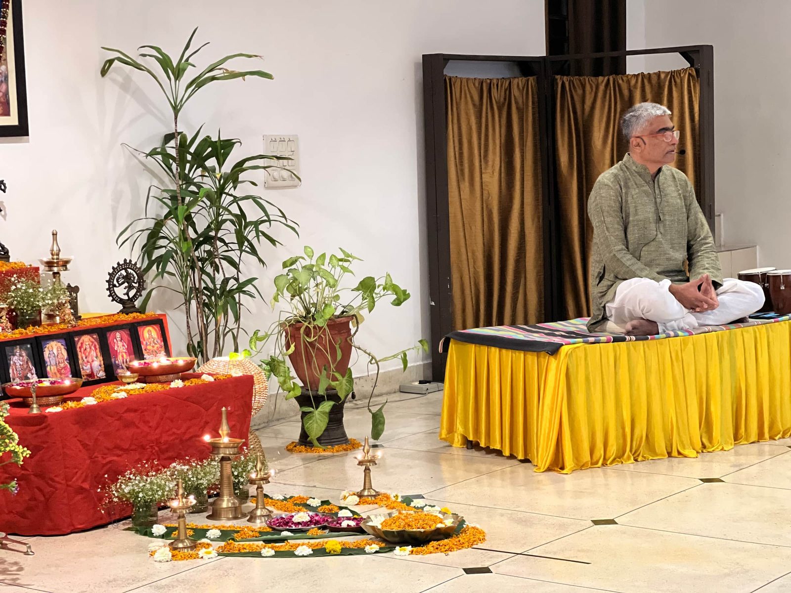A teacher seated cross-legged on a draped platform beside an altar of marigolds, oil lamps and framed images