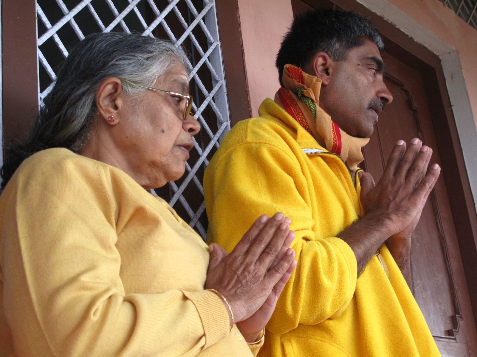 An older woman and a man in yellow, both with palms together in namaskar, photographed in profile