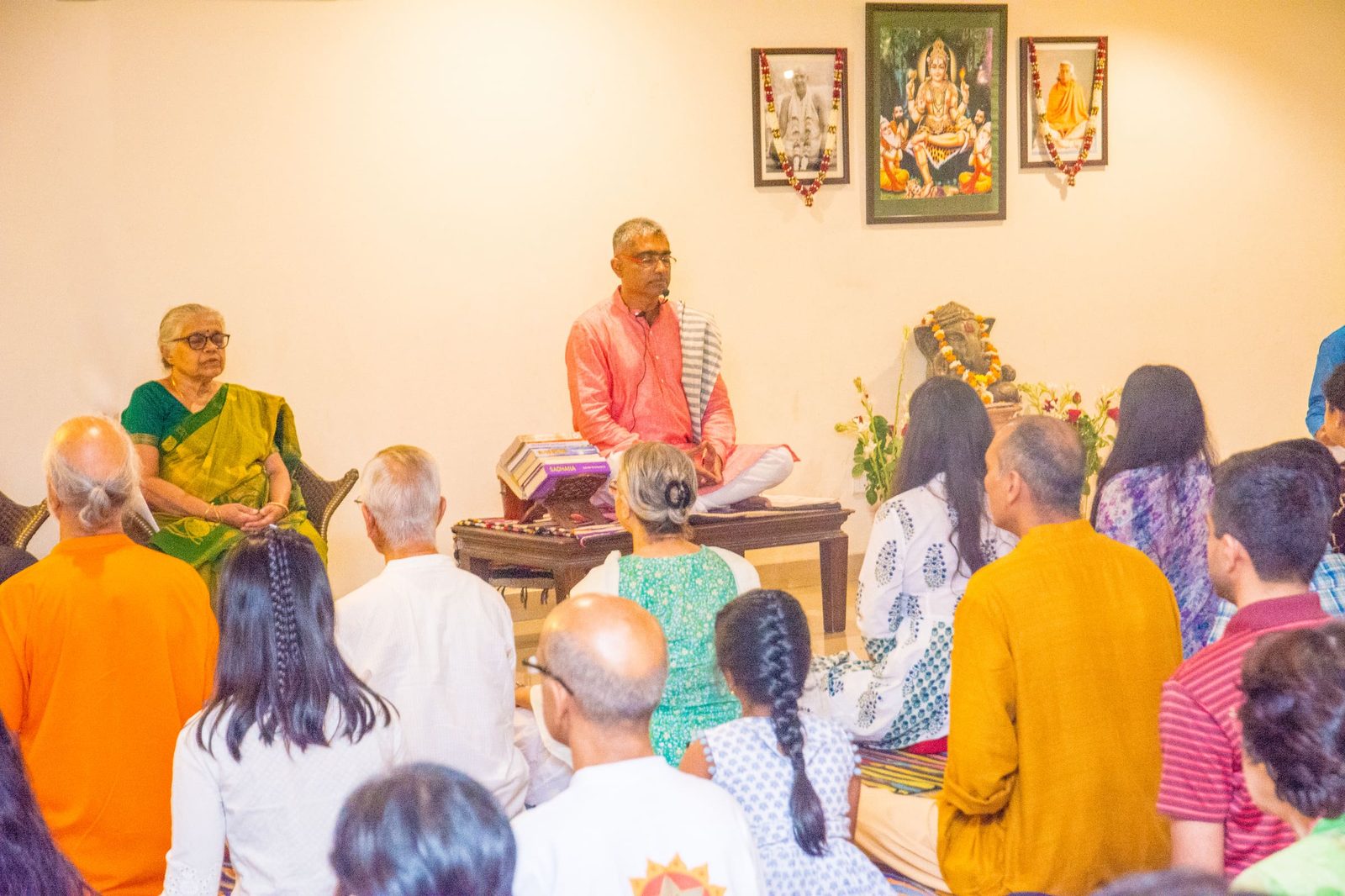A satsang, the teacher seated cross-legged at the front and the room seated facing him, framed portraits and a garlanded image behind