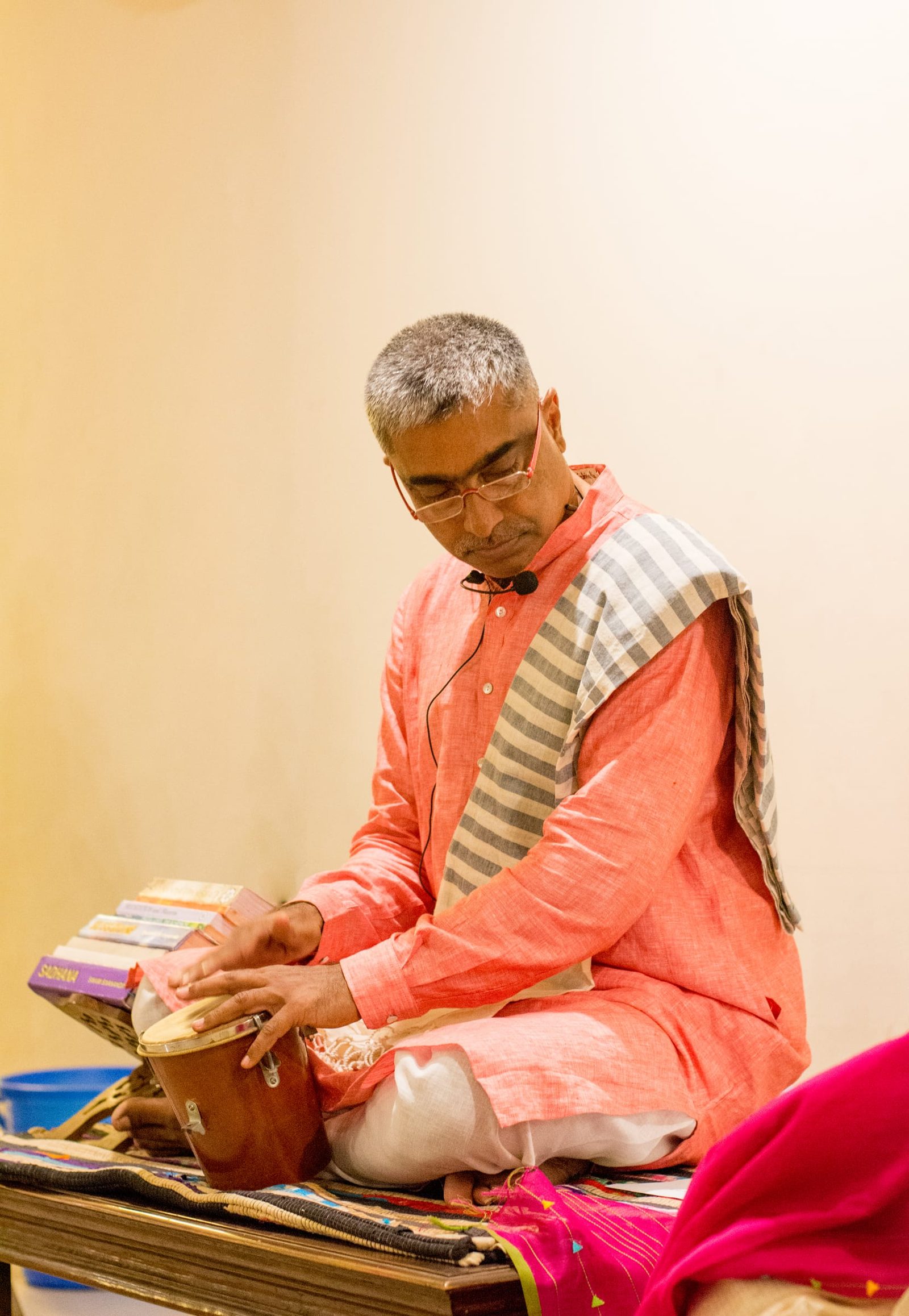A teacher seated in a peach kurta playing a hand drum, a stack of books beside him