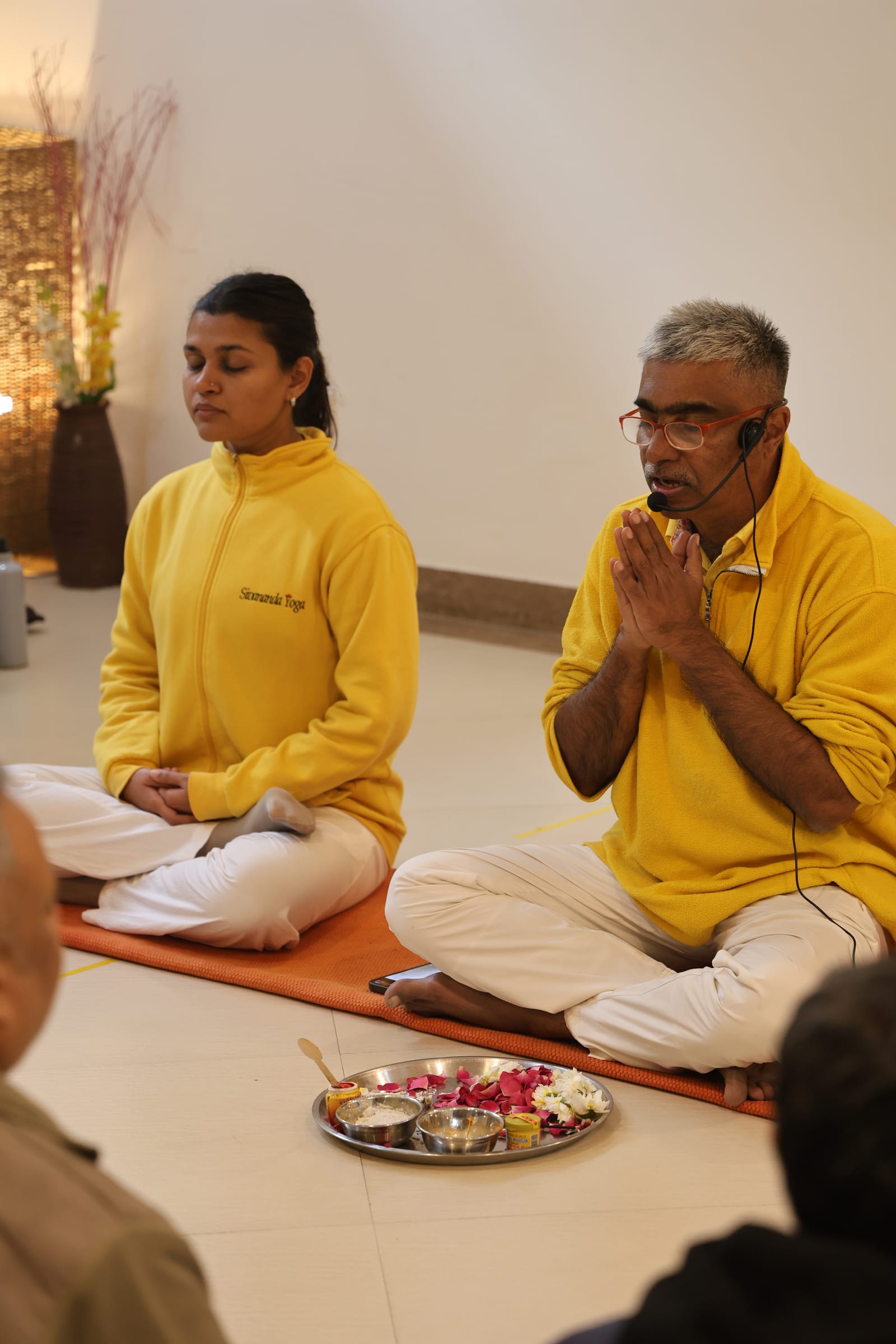 Two teachers in yellow fleeces seated cross-legged on a mat, the man on the right with palms together, a thali of rose petals on the floor before them