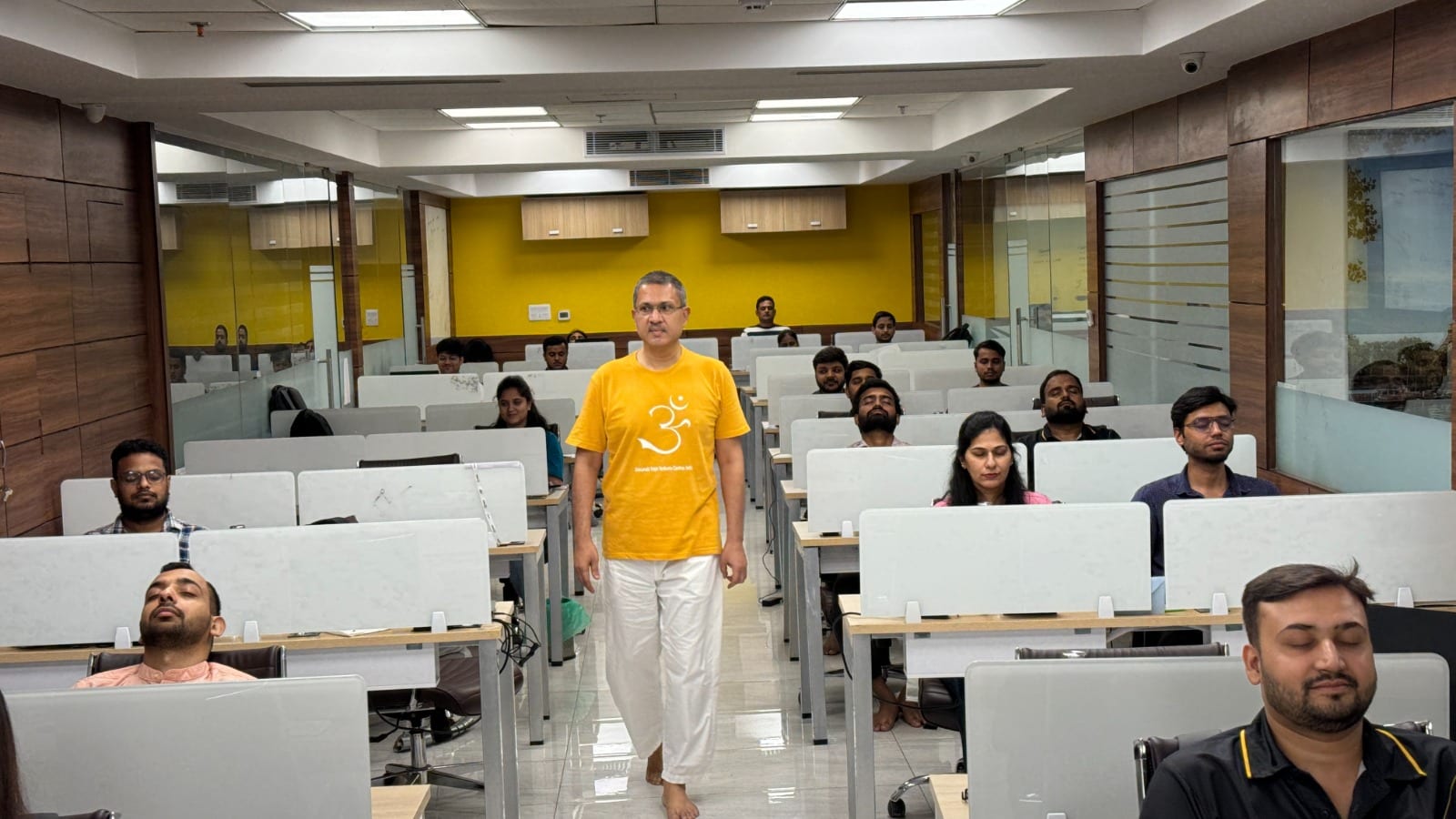 A teacher walking the aisle of an open-plan office while staff sit at their desks with eyes closed