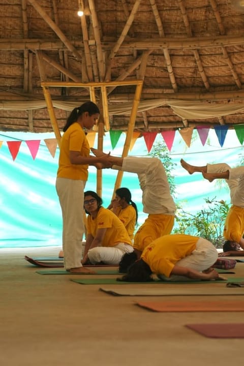 A Sivananda Gurgaon teacher training class: a teacher assisting a trainee into an inverted posture while other trainees practice in the thatched hall.