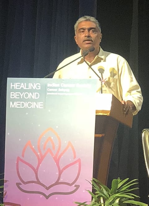 Yogacharya Arun Pandala speaking from a lectern, a banner for the Indian Cancer Society's Healing Beyond Medicine event standing in front of him.