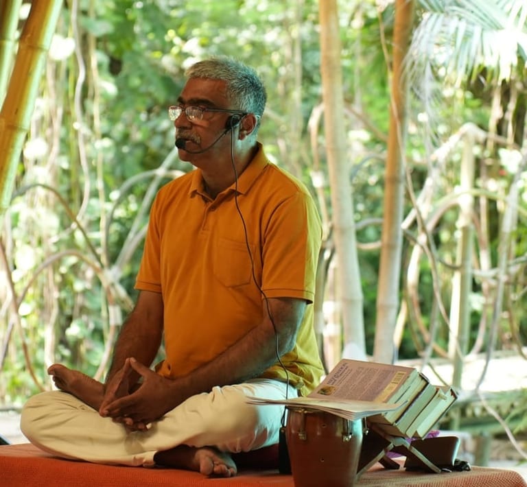 Arun Pandala speaking at Sivananda Gurgaon, seated cross-legged with a headset microphone, beside open books on a stand.