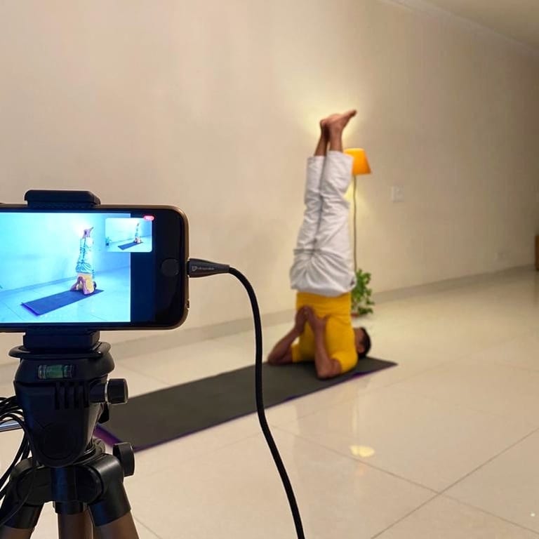 A teacher at Sivananda Gurgaon in shoulderstand on a mat at home, filmed on a phone mounted on a tripod for a class streaming live on Zoom.