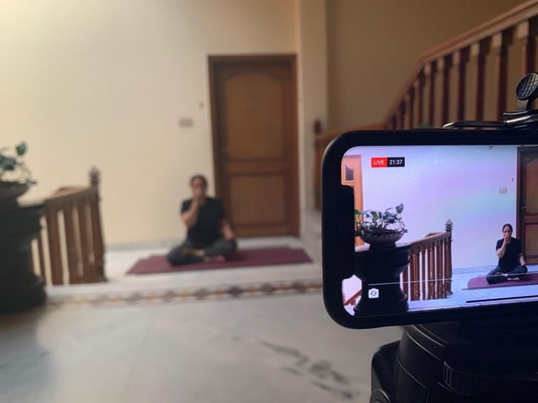 A phone on a tripod filming a live class, the teacher seated on her mat at the foot of a staircase and framed on its screen.