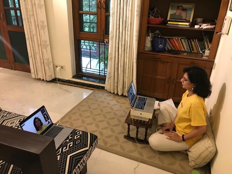 A student sitting cross-legged on the floor at home in front of her laptop, the teacher on a second screen beside her.