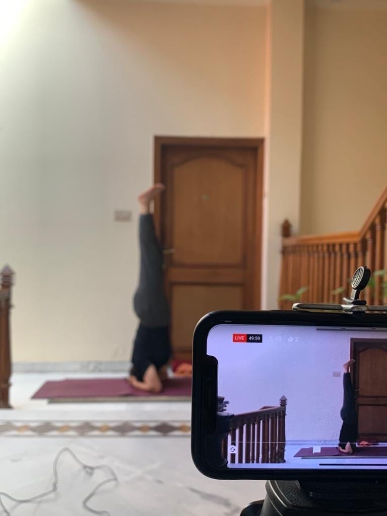 A phone on a tripod carrying a live class, a student in Headstand in the room behind it.