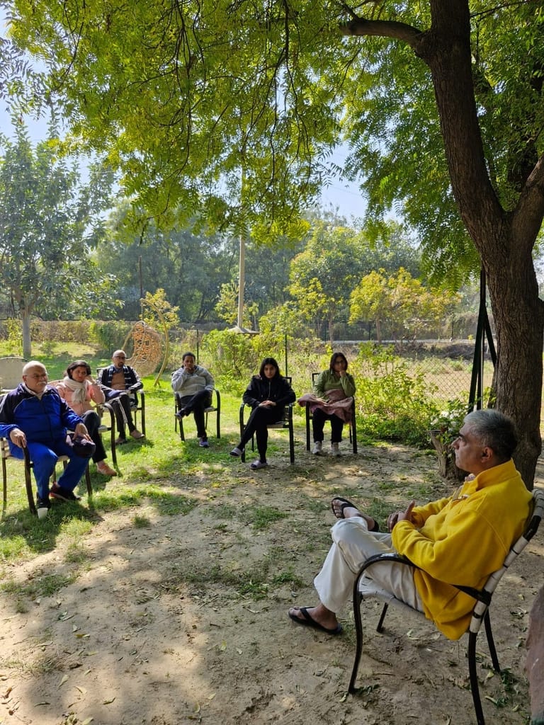 A group seated in a circle of chairs on grass under a tree, a teacher in a yellow fleece listening from the right