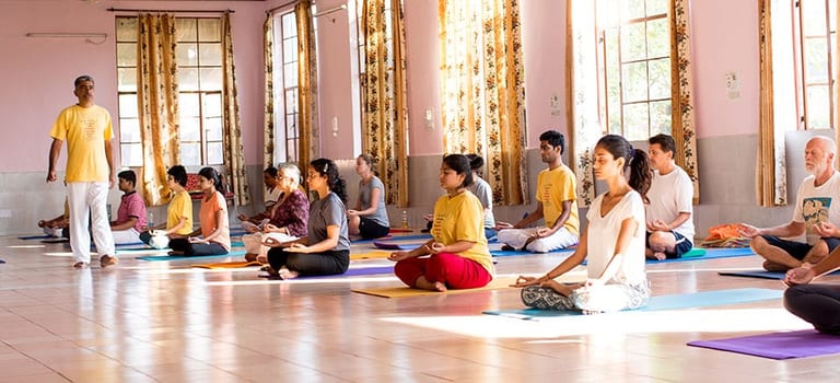A hall of students seated on mats in meditation, a teacher in a yellow shirt standing at the left