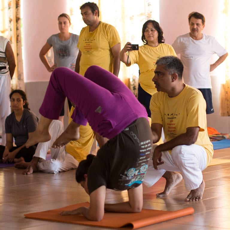 A teacher crouching beside a student on an orange mat as she tucks her head down towards a headstand, others watching